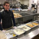 Person stands and smiles in front of trays of food in an industrial kitchen