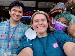 Three people stand smiling in front of a t-rex head: man with blue plaid shirt, woman in a brown beanie, and a woman with blue hair giving a thumbs-up.