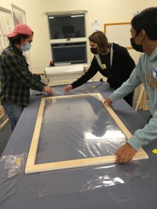 Three students hold a wooden frame flat on a table as they wrap it in clear plastic.
