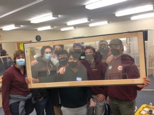 Group of masked students peer through a window insert made of pine and wrapped in clear plastic 