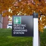 Image of sign that says "Electric Vehicle Charging Station" with bright orange tree in background