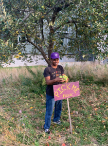 Student in purple Williams baseball cap stands behind a handmade sign with text saying "pick me please" and holds a handful of apples