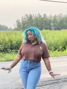 Micaela Foreman poses smiling in front of a field, with blue hair, a brown shirt and jeans