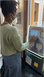 Woman wearing mask uses touchscreen on box to receive GenCo products