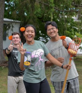 Three students stand outdoors with flowers behind their ears smile and give a thumbs up and the peace sign