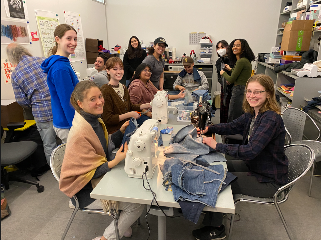 Group of students sit and stand around a table with sewing machines and denim