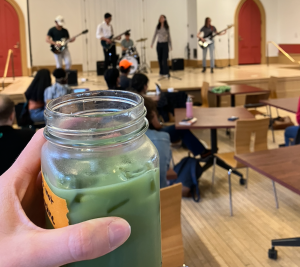 Mason jar mug filled with iced matcha is held up in front of a stage with performers blurred in the background