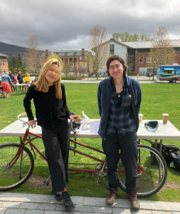 Two students stand smiling as they lean against a double-bicycle