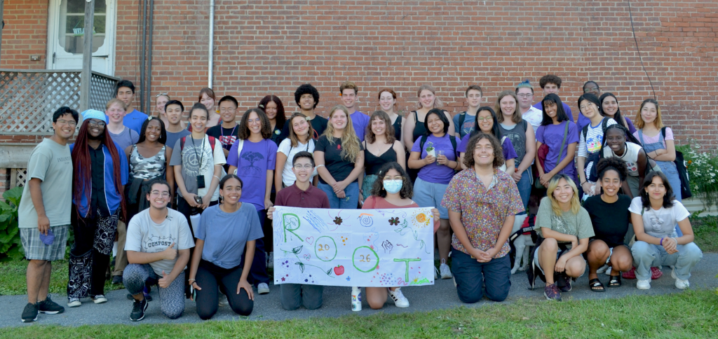 Large group of students stand smiling in front of a brick wall holding a cloth sign that says "Root"