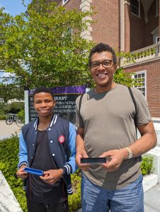 Milton and Chris stand smiling outside a brick building holding a 3-D printed part