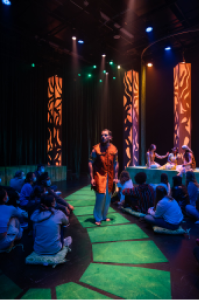 Actor in dark room walking on green floor tile pathway, surrounded by audience members sitting on the ground