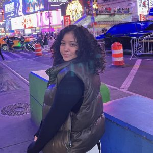 Woman in curly hair facing the camera wearing a gray puffer vest over a black long sleeve. She is centered in the middle of a busy, lit-up street at night.