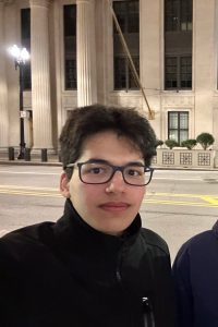 Man in front of a road. He has dark brown hair, rectangle glasses and is wearing a black turtleneck.