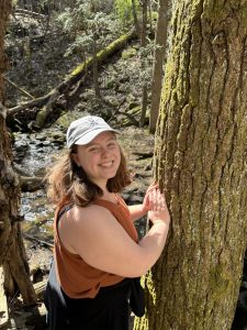 Person wearing a white cap and a brown tank top, leaning against a tree. They are in front of a stream on a sunny day.