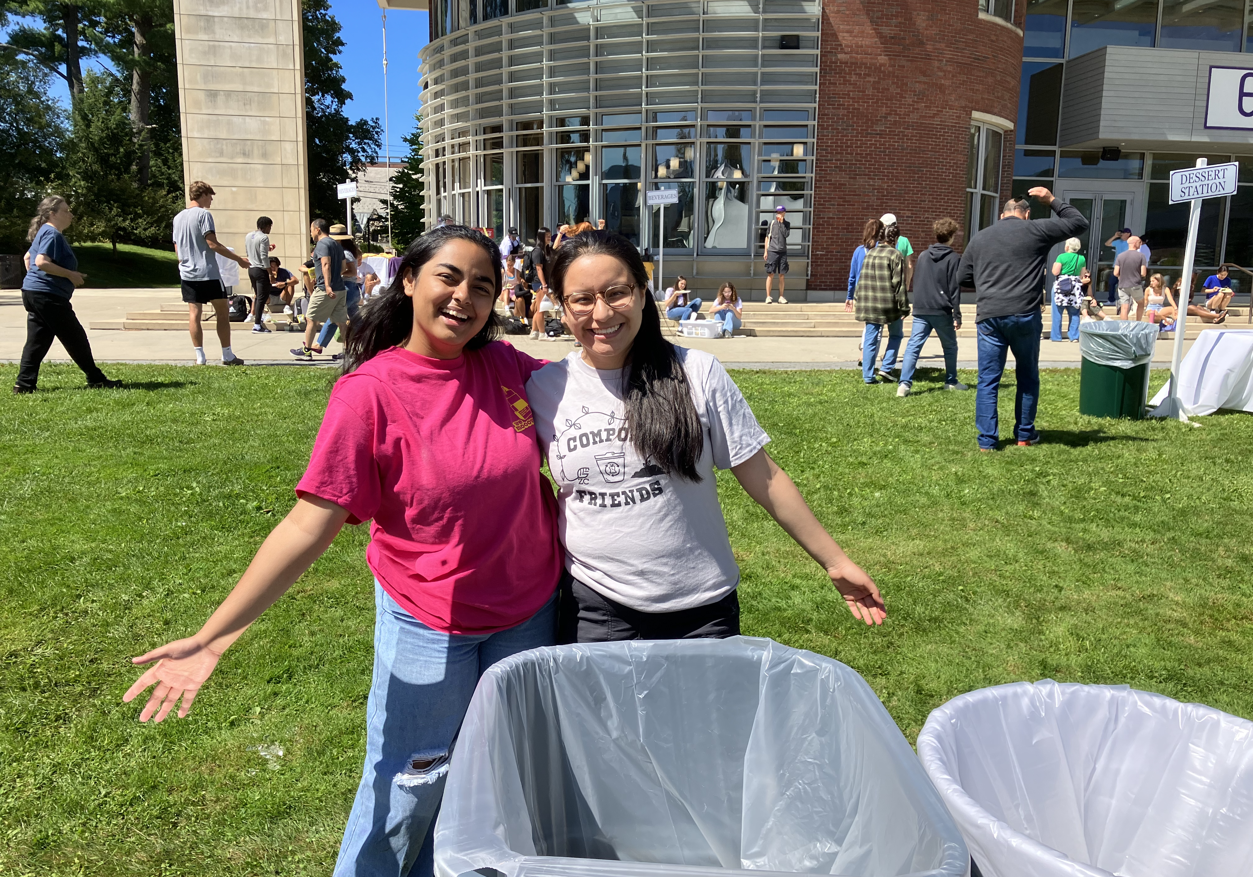Two students with long dark hair, one in a hot pink shirt and one in a grey "Compost Friends" shirt stand smiling next to a compost bin outdoors at an event