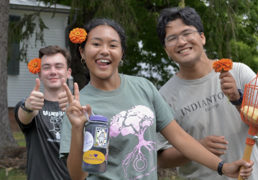 Three people stand smiling, with orange flowers behind their ears, giving thumbs up and the peace sign